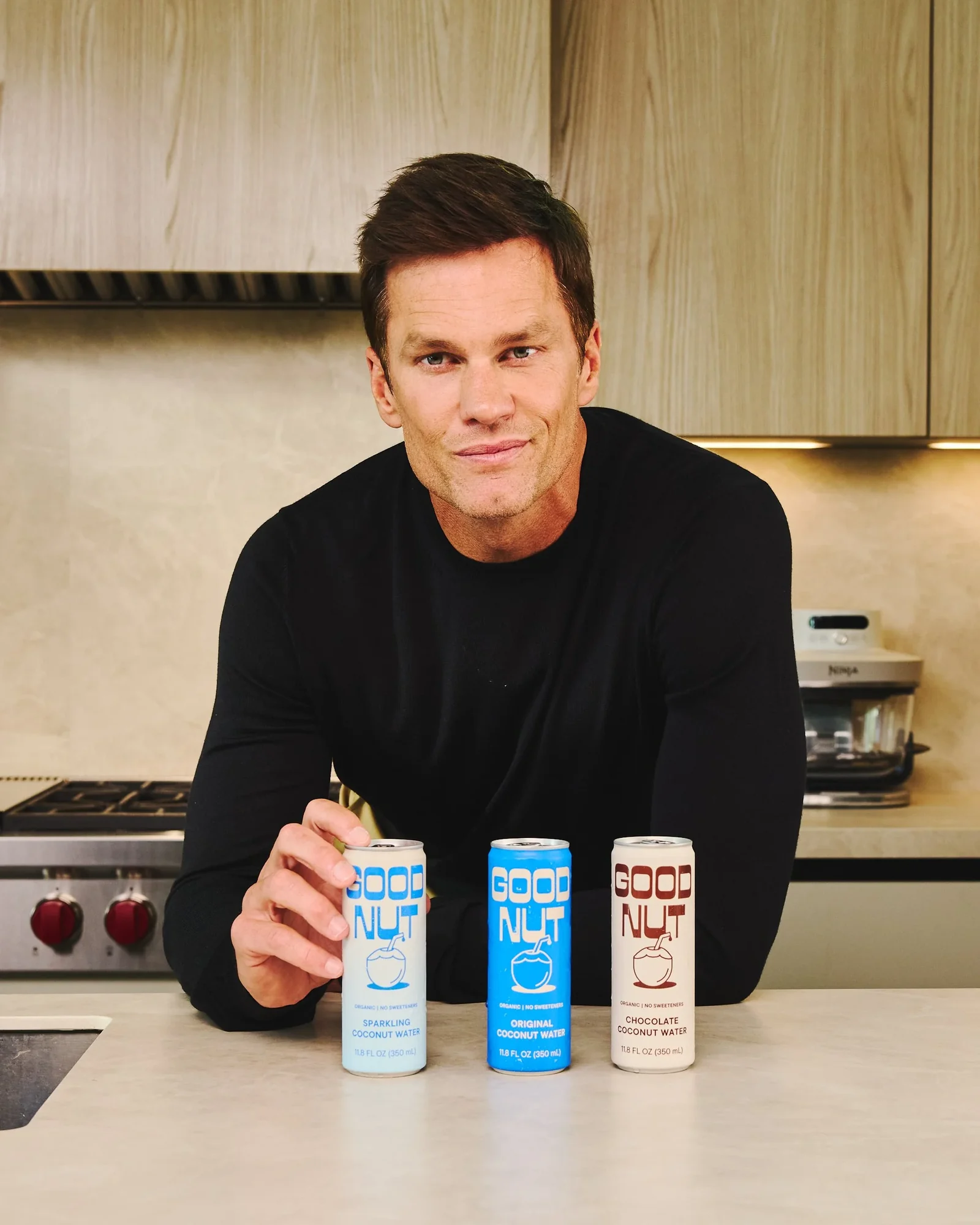 Tom Brady standing in a kitchen behind three cans of GOOD NUT coconut water